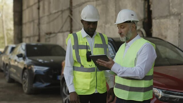 Two Construction Workers In Hardhats Looking At Tablet Screen While Standing Outdoor With Automobiles On Background. Multiethnic Men Talking About New Architect Ideas On Working Project.