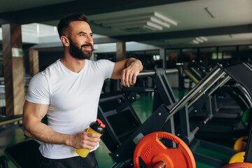 Lifestyle portrait of handsome muscular man standing with shaker after the training in the sport gym