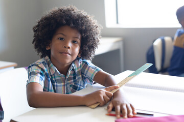 Portrait of african american elementary schoolboy holding ruler while sitting at desk in classroom
