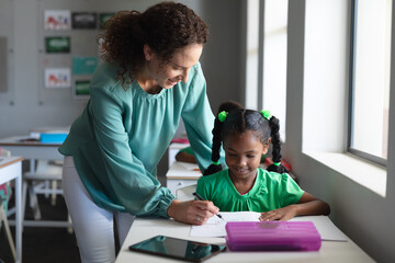 Caucasian young female teacher assisting african american elementary schoolgirl writing on book