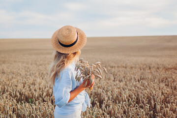 Back view of young woman walking in summer field wearing straw hat and linen shirt holding wheat...