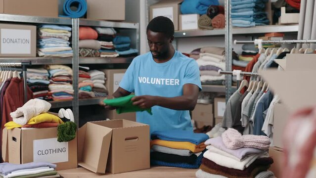 African american volunteer working at the warehouse while sorting and iterating clothes for donations. Belongings at the shelves at the background. Humanitarian aid concept