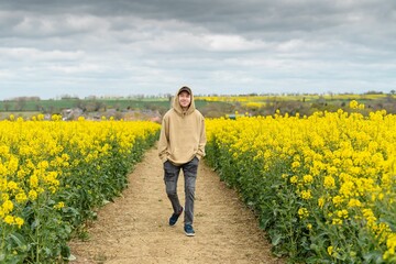 Young Caucasian man in a hoodie walking on a pathway through the white mustard field - Powered by Adobe