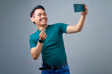 Smiling young Asian man taking selfie on mobile phone, showing Korean heart with two fingers...