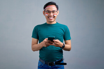 Smiling young Asian man in casual t-shirt  holding mobile phone and looking at camera isolated on grey background. indonesian independence day celebration concept