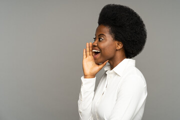 Successful black businesswoman with kinky hair holds hand near mouth speaking. Young African American woman in classic white shirt says advertising text in empty space standing on grey background