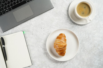 A croissant and a cup of coffee on white plates on grey working desk in the office with laptop and notebook, copy space for text