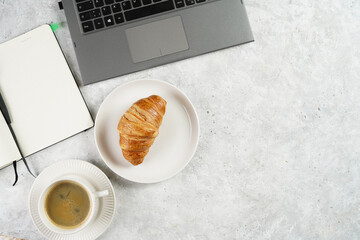 A croissant and a cup of coffee on white plates on grey working desk in the office with laptop and notebook, copy space for text