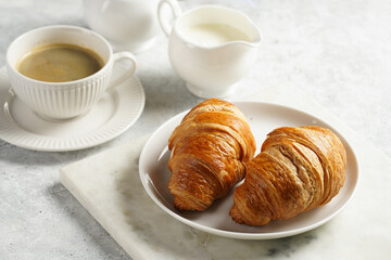 Two french soft croissants on a white plate on a marble tray, a cup of black coffee in white sophisticated cup, milk, sugar, jam