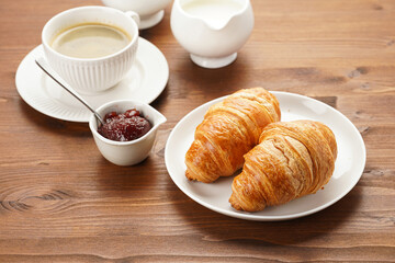 Two french soft croissants on a white plate on a wooden table, a cup of black coffee in white sophisticated cup, milk, sugar, jam