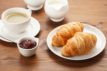 Two french soft croissants on a white plate on a wooden table, a cup of black coffee in white sophisticated cup, milk, sugar, jam