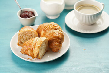 Two french soft croissants on a white plate on a blue wooden table, a cup of black coffee in white sophisticated cup, milk, sugar, jam