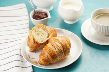 Two french soft croissants on a white plate on a blue wooden table, a cup of black coffee in white sophisticated cup, milk, sugar, jam