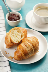 Two french soft croissants on a white plate on a blue wooden table, a cup of black coffee in white sophisticated cup, milk, sugar, jam