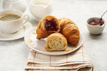 Two french soft croissants with jam on a white plate on a checkered napkin, a cup of black coffee in white sophisticated cup, milk, sugar