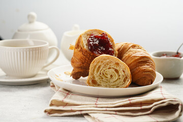 Two french soft croissants with jam on a white plate on a checkered napkin, a cup of black coffee in white sophisticated cup, milk, sugar