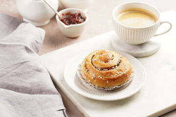 A round cinnamon pastry on a white plate on marble board, cup of coffee, jam, milk