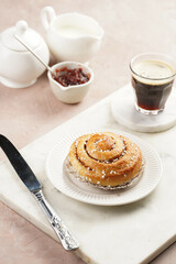 A round cinnamon pastry on a white plate on marble board on light pink background, cup of coffee, jam, milk