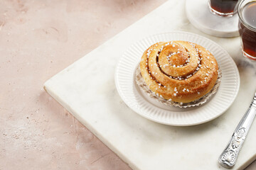 A round cinnamon pastry on a white plate on marble board on light pink background, cup of coffee, jam, milk