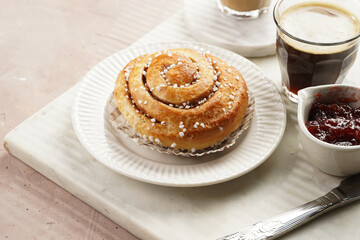 A round cinnamon pastry on a white plate on marble board on light pink background, cup of coffee, jam, milk
