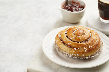 A round cinnamon pastry on a white plate on marble board on light pink grey, cup of coffee, jam, milk