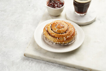 A round cinnamon pastry on a white plate on marble board on light pink grey, cup of coffee, jam, milk