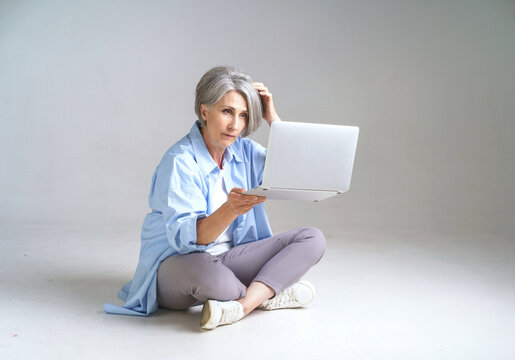 Frustrated Mature Woman Holding Laptop In Hand Scratching Her Head Looking At It Sitting On The Floor Isolated On White Background. Mature People And Technologies. Senior Woman With Laptop.