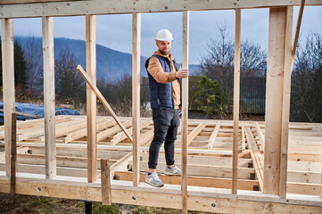Male builder building wooden frame house. Portrait of man standing on construction site in safety helmet, inspecting quality of work.