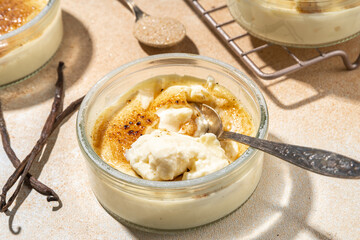 Sweet creme brulee dessert. Portioned glass jars with homemade creme brulee with caramel topping, on beige colored background with sugar in spoon and vanilla copy space