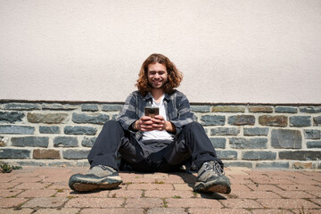 Fototapeta premium A happy young Caucasian guy sitting in his phone against the wall