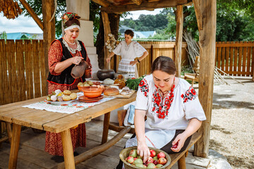 A cheerful company of village women in national Ukrainian Kuban Russian Belarusian clothes. Kuban Cossacks. Cooking traditional food in the summer kitchen in the courtyard - obrazy, fototapety, plakaty