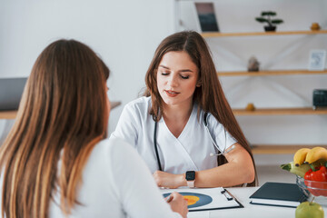 Female visitor is sitting with doctor. Professional medical worker in white coat is in the office