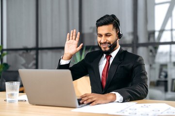 Positive successful indian or arabian man in suit, with headset, business advisor, call center worker, company representative, support service operator, holds online consultation, talking with client