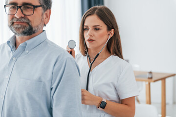Woman using stethoscope and listening lungs of the man. Professional medical worker in white coat is in the office