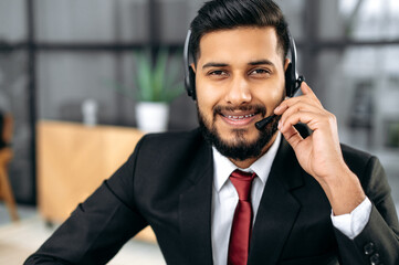 Portrait of friendly positive smart indian or arabian man with headset, call center worker, expert, company representative, support service operator, holds online consultation, looks at camera, smiles