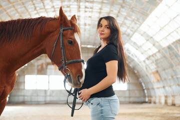 Looking at the camera. Beautiful pregnant woman in casual clothes is with the horse on a stable