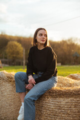 Naklejka na ściany i meble Photo of a beautiful woman in casual clothes sitting on a hay bale and looking away with a serious face. Vertical