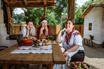 three cheerful village women in national Ukrainian Kuban Russian Belarusian clothes are sitting chatting on a summer evening near a village house. Friendly communication in the company at the common t - obrazy, fototapety, plakaty