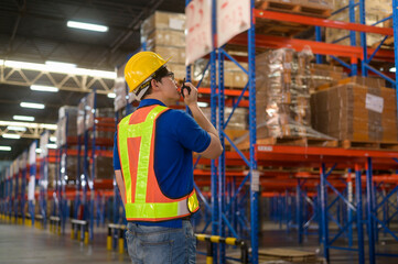 Young asian male worker wearing helmet using talki walki in modern warehouse.