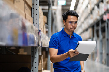 Asian warehouse worker with clipboard inspecting parcel boxes in warehouse warehouse. Analysis of new arrivals for placement in storage department. Work at the warehouse.