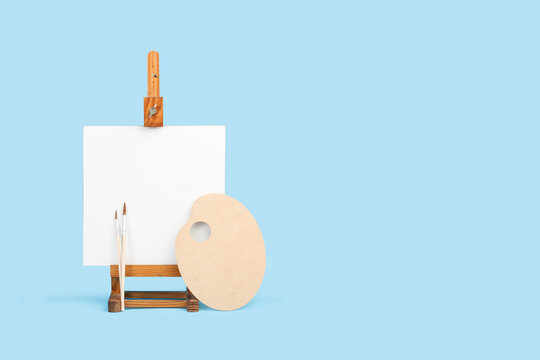 Lienzo en blanco con caballete de madera junto a una paleta y pinceles sobre un fondo celeste claro liso y aislado. Vista de frente y de cerca. Copy space