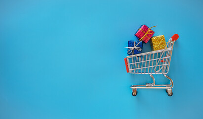 Flat lay view of small shopping cart and gift box on blue background with copy space 