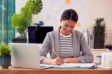 Female Architect Working In Office Sitting At Desk Studying Plans For New Building