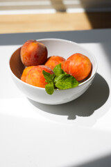 Tasty healthy snacks, white bowl with ripe fruit, fresh peaches,  mint leaf and glass on light table in daylight