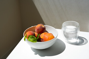 Close-up of fresh peaches in a white bowl with a sprig of mint standing on a table in daylight next to a glass of water.