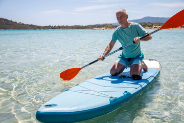 Sup surfing. Beautiful view of the sea with a mature man kneeling on a board with paddle in the water. Standup paddleboarding in summer