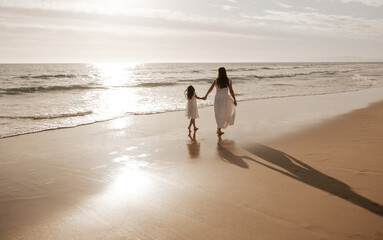 mother with daughter holding hands and walking on beach at sundown