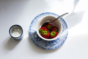 Selective focus high angle view of strawberries in plain white bowl set on pretty old plate with blue pattern, with antique sugar bowl next to it 