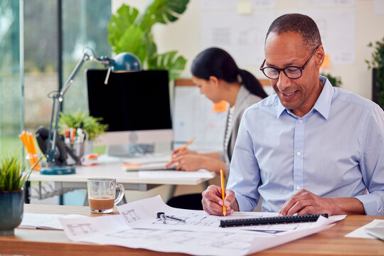 Male And Female Architects Working In Office At Desks On Plans For New Building