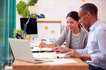 Male And Female Architects Working Together In Office At Desks On Plans For New Building